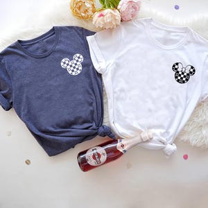 May include: Two t-shirts with a checkered Mickey Mouse design. The shirt on the left is navy blue and the shirt on the right is white.