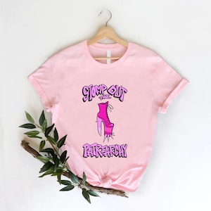 May include: A light pink t-shirt with the words "STOMP OUT THE PATRIARCHY" in purple and a pink high-heeled boot graphic. The shirt is hanging on a wooden hanger.