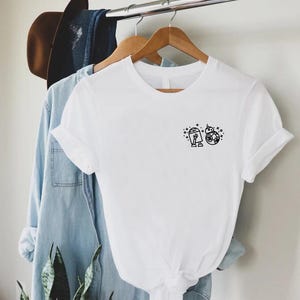 May include: White t-shirt with a small black graphic of two Star Wars droids, R2-D2 and BB-8, on the chest. The shirt is hanging on a wooden hanger with other clothing items, including a denim shirt and a brown hat.