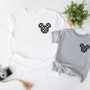 May include: Two t-shirts, one white and one gray, with a black and white checkered Mickey Mouse head design on the front.