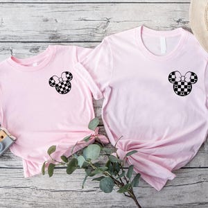 May include: Two pink t-shirts with a black and white checkered Mickey Mouse head design on the front. The shirts are laid out on a wooden surface.