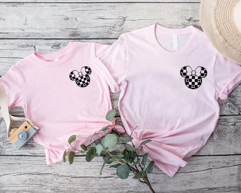 May include: Two pink t-shirts with a black and white checkered Mickey Mouse head design on the front. The shirts are laid out on a wooden surface.