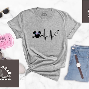 May include: A grey t-shirt featuring a black and white graphic of a Mickey Mouse head with flowers on top and a stethoscope. The graphic is next to a black and white heartbeat line. The t-shirt is on a white surface with a pair of sunglasses, a pink sign that says "Ships Today", a pair of jeans, a watch, and a brown tag that says "Mama Bear" with a bear logo.