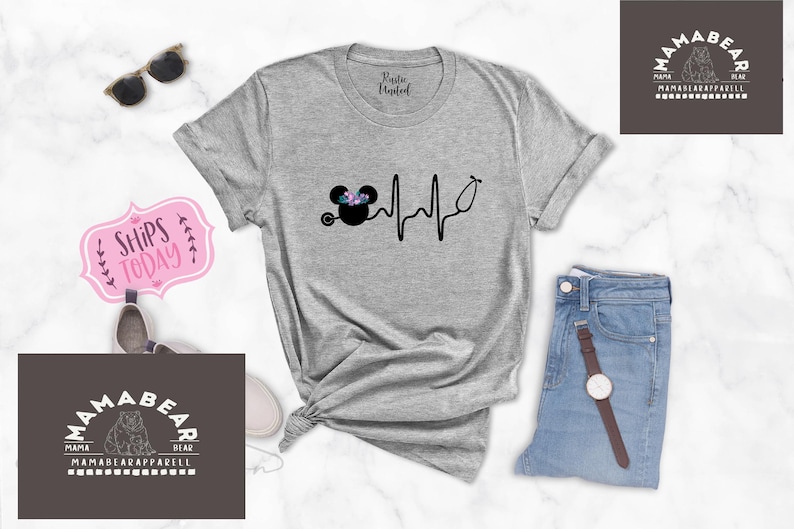 May include: A grey t-shirt featuring a black and white graphic of a Mickey Mouse head with flowers on top and a stethoscope. The graphic is next to a black and white heartbeat line. The t-shirt is on a white surface with a pair of sunglasses, a pink sign that says "Ships Today", a pair of jeans, a watch, and a brown tag that says "Mama Bear" with a bear logo.