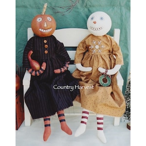 May include: Two handmade rag dolls sitting on a white bench. The doll on the left has a pumpkin head and is wearing a black and white striped dress. The doll on the right has a white head and is wearing a brown dress with white polka dots and a snowflake design. The text "Country Harvest" is visible on the bench.