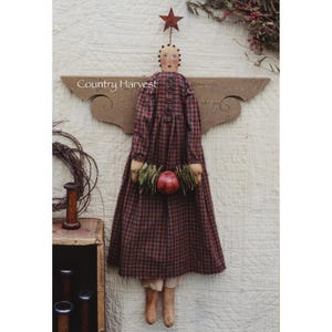May include: A handmade primitive style angel doll wearing a red and black checked dress, holding a wreath of greenery and a red apple. The doll has a star on its head and is standing on a wooden shelf with the text "Country Harvest" on it.