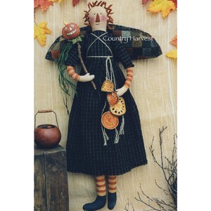 May include: A handmade rag doll dressed in a black and white plaid dress with orange and red striped stockings. The doll is holding a stick with a jack-o-lantern on top and a string of dried orange slices. The doll is standing in front of a brown background with the text "Country Harvest" on a plaid fabric.