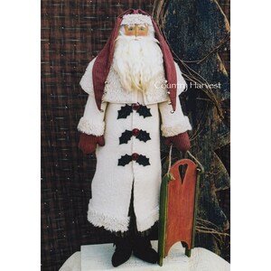 May include: A white-bearded Santa Claus doll wearing a white fur coat with black holly berry buttons, a red hat, and red mittens. He is holding a red wooden sled with a green base. The doll is standing in front of a wooden background with the text "Country Harvest".