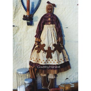 May include: A brown gingerbread doll wearing a red and white checked dress and a brown apron. The doll is holding three gingerbread people in her hands. The doll is standing on a brown surface with cinnamon sticks and jars in the background. The text "Country Harvest" is printed on the doll's apron.