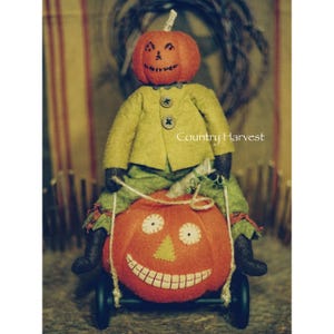 May include: A handmade felt pumpkin doll with a green jacket and black pants riding a pumpkin on wheels. The pumpkin has a smiling face with white eyes and a stitched mouth. The doll's head is also a pumpkin with a carved face. The text "Country Harvest" is visible in the image.