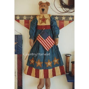 Könnte beinhalten: Ein brauner Teddybär, der ein blau-weiß gestreiftes Kleid mit roten, weißen und blauen Sternen und einer rot-weiß-blauen Flagge trägt. Der Bär hält einen Stock mit einer kleinen amerikanischen Flagge daran. Der Text "Country Harvest" ist unter dem Bären zu sehen.