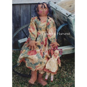 May include: Two rag dolls, one large and one small, are sitting in a wooden wagon. The larger doll has brown skin, black hair, and is wearing a floral dress. The smaller doll has white skin, red hair, and is wearing a red and white gingham dress. The wagon is painted blue and green.