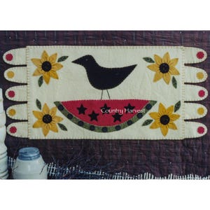 May include: A felt wall hanging featuring a black bird perched on a slice of watermelon. The watermelon is red with black stars and a green rind. There are yellow sunflowers on either side of the bird and watermelon. The wall hanging has a white background and a brown border. The text "Country Harvest" is printed on the watermelon.
