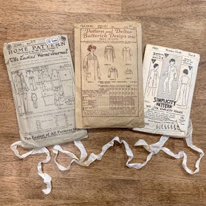 May include: Three vintage sewing pattern packets. The patterns are for clothing, including a slumber outfit and other garments. The packets are tan and cream-colored with illustrations and text. White ribbon is draped across the bottom of the packets.