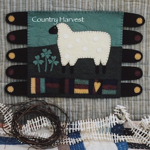 Puede incluir: Un tapiz de pared de fieltro con el texto "Country Harvest" presenta una oveja blanca con manchas negras de pie en un campo verde con tres tréboles. La oveja está de pie sobre una valla marrón con un corazón rojo.