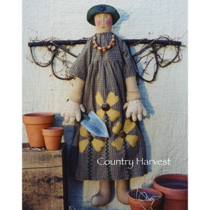 May include: A handmade rag doll dressed in a plaid dress with yellow flower accents. The doll is holding a small gardening tool and is standing in front of a wooden crate with terracotta flower pots. The doll's wings are made of twigs and vines. The text "Country Harvest" is printed below the doll.
