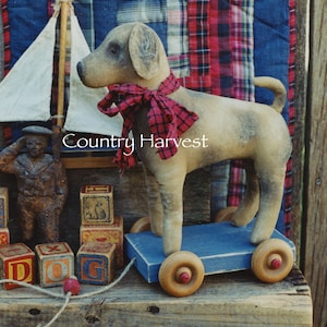 Peut inclure: Un jouet en forme de chien beige avec une écharpe à carreaux rouges, monté sur un chariot en bois bleu avec des roues en bois. L'image comprend le texte "Country Harvest", une figurine de marin et des blocs alphabet.