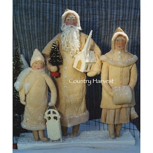 Op de afbeelding: Een vintage kerstscène met een kerstmanpop, een meisjespop en een kleine kerk. De poppen zijn gekleed in wit en hebben een besneeuwde, winterse uitstraling. De tekst "Country Harvest" is zichtbaar in de afbeelding.