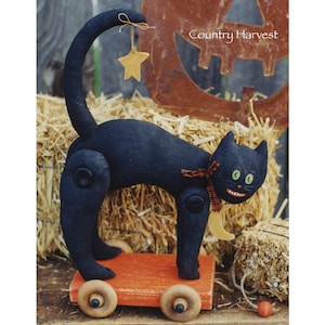 Könnte beinhalten: Eine schwarze Katze mit einem grinsenden Gesicht und grünen Augen sitzt auf einem roten Holzwagen mit Rädern. Die Katze trägt ein braunes und weißes kariertes Band um den Hals und einen gelben Halbmond auf der Brust. Der Schwanz der Katze ist eingerollt und mit einer Schnur an einem gelben Stern befestigt.