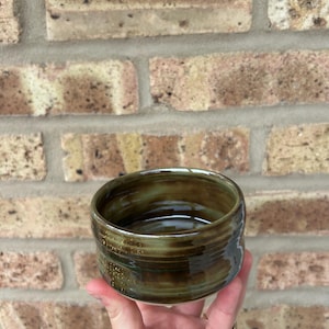 May include: A small, dark green ceramic bowl with a brown glaze and gold flecks. The bowl has a rounded shape and a slightly flared rim.