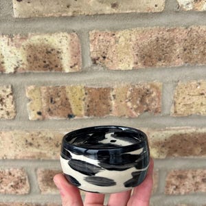 May include: A small, black and white ceramic bowl with a glossy finish. The bowl has a unique abstract pattern.