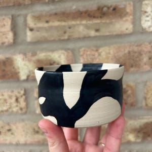 May include: A small, white ceramic bowl with black abstract designs. The bowl is hand-painted and has a slightly irregular shape.