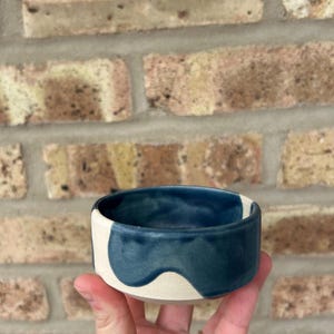 May include: A small, white ceramic bowl with a blue glaze. The glaze has a wavy, abstract design.