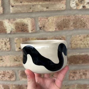 May include: A small, white ceramic bowl with a black, abstract design. The bowl has a rounded shape and a smooth, glossy finish.