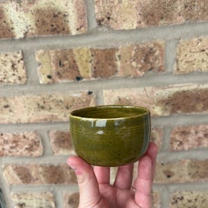 May include: A small, green ceramic bowl with a speckled glaze. The bowl is round and has a slightly flared lip.