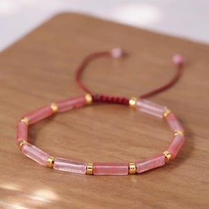 Peut inclure: Un bracelet en perles rose et or avec une fermeture en cordon rouge. Le bracelet est composé de petites perles rectangulaires avec des accents dorés.