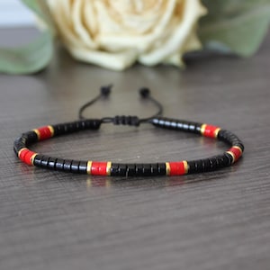 Op de afbeelding: Zwart kralenarmband met rode en gouden accenten. De armband heeft een verstelbare schuifknoopsluiting. De kleine, cilindrische kralen creëren een minimalistisch ontwerp. De armband wordt gepresenteerd op een donkergrijze ondergrond.