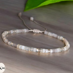 Puede incluir: Un brazalete de cuentas blancas y grises con cierre de nudo deslizante. El brazalete está hecho de pequeñas cuentas redondas con un brillo sutil.