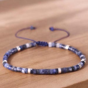 May include: A blue and silver beaded bracelet with a sliding knot closure. The bracelet is made with small, square-shaped beads.