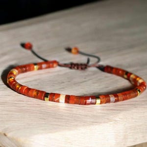 May include: A red and gold beaded bracelet with a sliding knot closure. The bracelet is made of small, square-shaped beads.