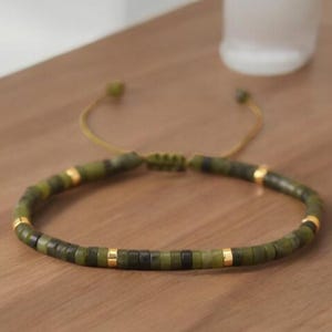 Pode incluir: Um bracelete de contas verdes e douradas com fecho de nó deslizante. O bracelete é feito com contas pequenas e redondas e espaçadores dourados.