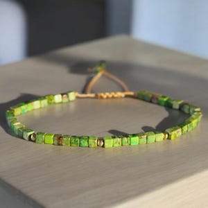 Könnte beinhalten: Ein Armband aus grünen Edelsteinperlen mit einem braunen Kordelverschluss. Die Perlen sind quadratisch und haben eine hellgrüne Farbe.