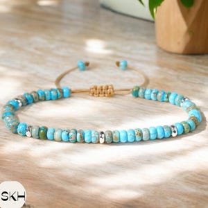 Puede incluir: Una pulsera de cuentas turquesas con detalles plateados y un cordón beige tejido. La pulsera presenta pequeñas cuentas redondas en tonos azules, verdes y grises. El cordón ajustable permite un ajuste cómodo.