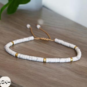 May include: White and gold beaded bracelet with a sliding knot closure. The beads are white with a marbled pattern and the knot is gold.