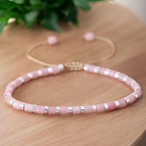 May include: A pink beaded bracelet with silver-coloured accents. The bracelet is adjustable with a thin, gold-coloured cord and small pink beads at the ends. The bracelet is displayed on a wooden surface.