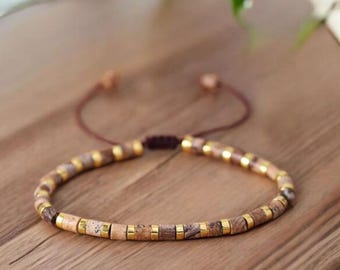 May include: A beaded bracelet featuring natural stone beads in shades of brown and tan, interspersed with gold-colored metal accents. The bracelet is adjustable with a brown cord and is displayed on a wooden surface.