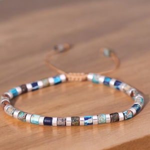 May include: A blue and silver beaded bracelet with a brown cord fastening. The bracelet is made of small, square beads in various shades of blue and silver.