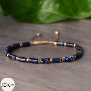 May include: Adjustable bracelet with dark blue and gold beads. The bracelet has small, rectangular beads with gold accents and a woven cord. The bracelet is displayed on a wooden surface.