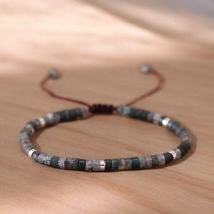 May include: A grey and silver beaded bracelet with a brown cord closure. The beads are small and round, and the cord is adjustable.