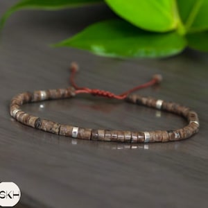 May include: A brown and silver beaded bracelet with a red cord closure. The bracelet is made of small, rectangular beads with a silver accent on each bead.
