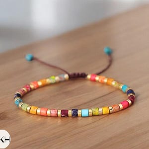 May include: A vibrant beaded bracelet featuring a spectrum of colours: red, orange, yellow, green, blue, and purple. The bracelet is strung on a brown cord with gold-coloured spacers. The bracelet is displayed on a wooden surface.