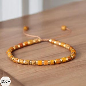 Puede incluir: Un brazalete de cuentas amarillas y doradas con cierre de nudo deslizante. El brazalete está hecho de pequeñas cuentas redondas con un borde dorado.