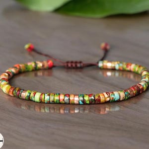 Può includere: Un braccialetto di perline colorate con chiusura in corda marrone. Le perline sono piccole e rotonde, e sono disposte in un motivo casuale. Il braccialetto è realizzato con materiali naturali e ha un aspetto rustico.