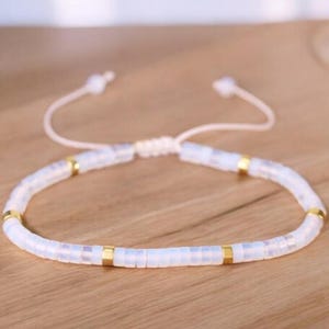 May include: A white beaded bracelet with gold accents. The bracelet is adjustable with a sliding knot.