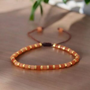 May include: A bracelet with orange and gold beads. The bracelet is strung on a brown cord with adjustable ends. The beads are small and cylindrical, with gold spacers. The bracelet is designed to be worn on the wrist. The bracelet's circumference is approximately 18 cm.