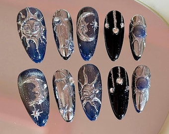 Puede incluir: Conjunto de diez uñas postizas con temática celestial. Las uñas están decoradas con lunas, soles y estrellas plateadas sobre un fondo azul y negro.